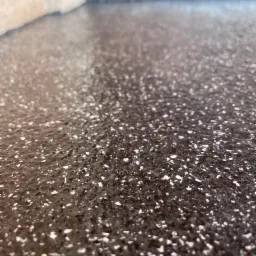 Błyszcząca, ciemna posadzka z drobnymi, jasnymi wtrąceniami, widoczna przy krawędzi ściany i progu drzwi.