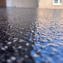 Woodstrug - Błyszcząca, ciemna posadzka żywiczna z delikatną strukturą, sfotografowana pod kątem, z rozmytym tłem budynku z oknem.