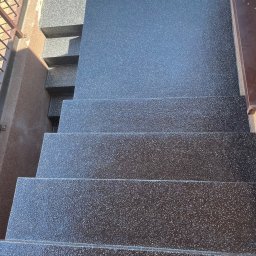 Woodstrug - Schody zewnętrzne pokryte ciemną, błyszczącą powłoką z widoczną fakturą, z metalową barierką po prawej stronie i fragmentem ceglanego muru w tle.