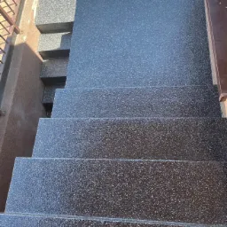 Schody zewnętrzne pokryte ciemną, błyszczącą powłoką z widoczną fakturą, z metalową barierką po prawej stronie i fragmentem ceglanego muru w tle.