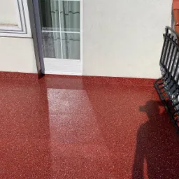 Balkon z czerwoną, błyszczącą posadzką z widoczną teksturą, cień osoby po prawej stronie, białe drzwi balkonowe i fragment metalowej balustrady.