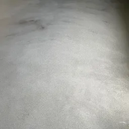 Szara, gładka powierzchnia podłogi z delikatnymi smugami i nierównomiernym rozkładem światła, przypominająca efekt polerowanego betonu.