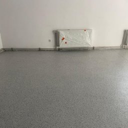 Woodstrug - Szara posadzka z drobnymi, ciemnymi plamkami, na ścianie grzejnik zabezpieczony folią. Widoczne rury grzewcze wzdłuż ściany. U góry zegar.