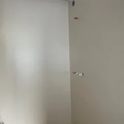 Beżowe ściany i podłoga w narożniku pomieszczenia z widocznymi elementami instalacji. Minimalistyczny design, gładkie powierzchnie, nowoczesne wykończenie.