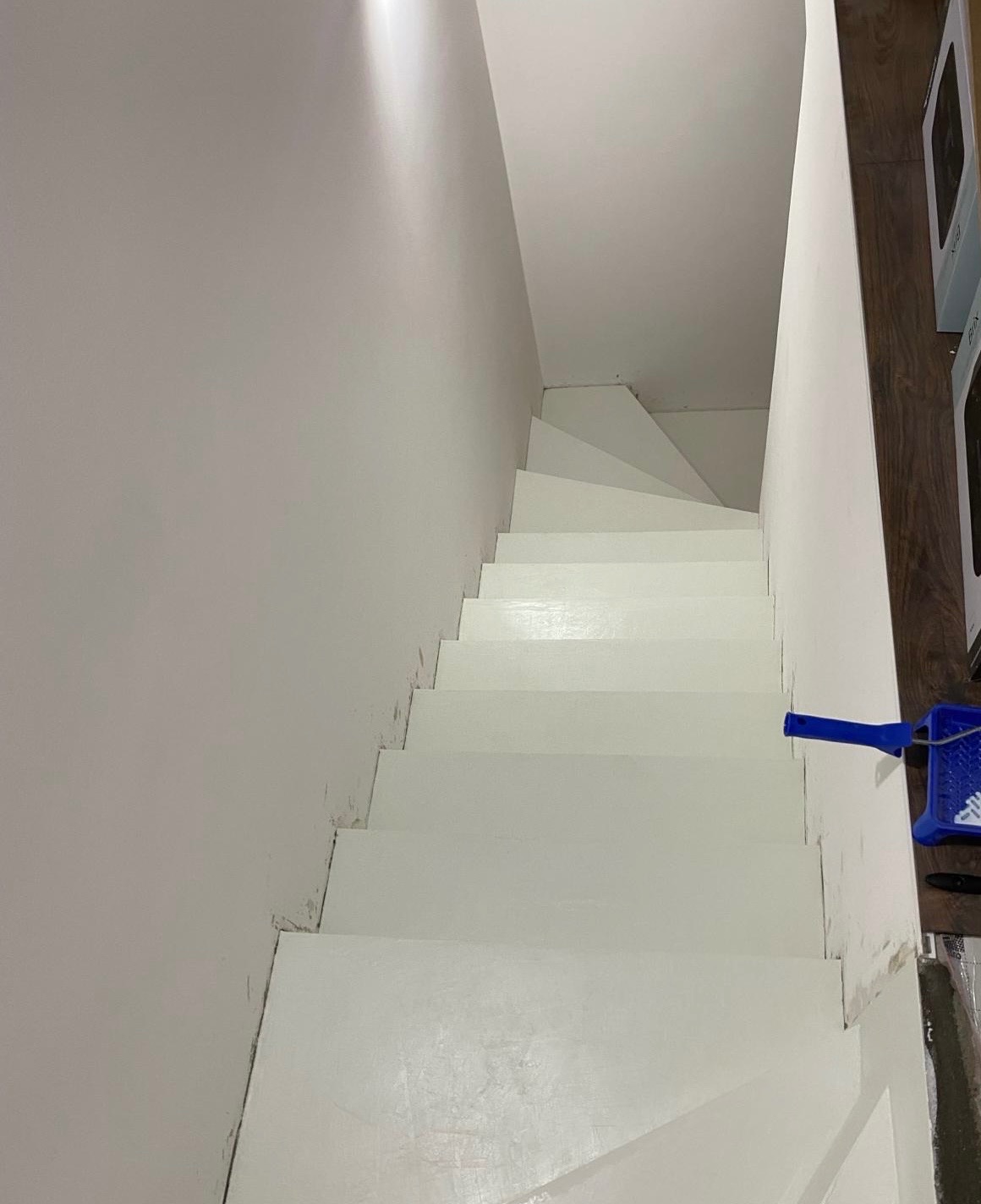 Białe schody pomalowane na biało, widoczny niebieski wałek do malowania i kuweta z farbą, ściany również białe.