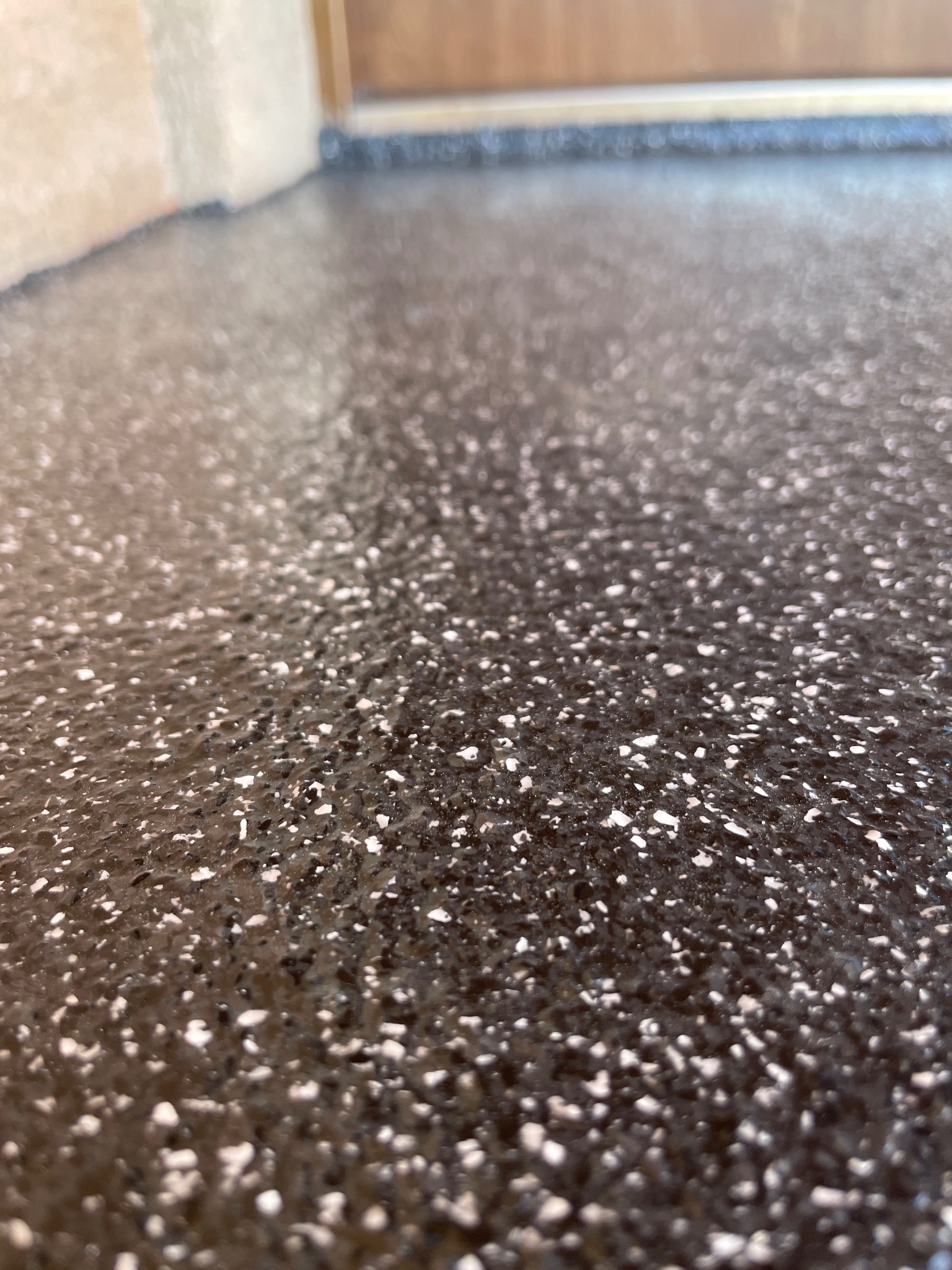 Błyszcząca, ciemna posadzka z drobnymi, jasnymi wtrąceniami, widoczna przy krawędzi ściany i progu drzwi.