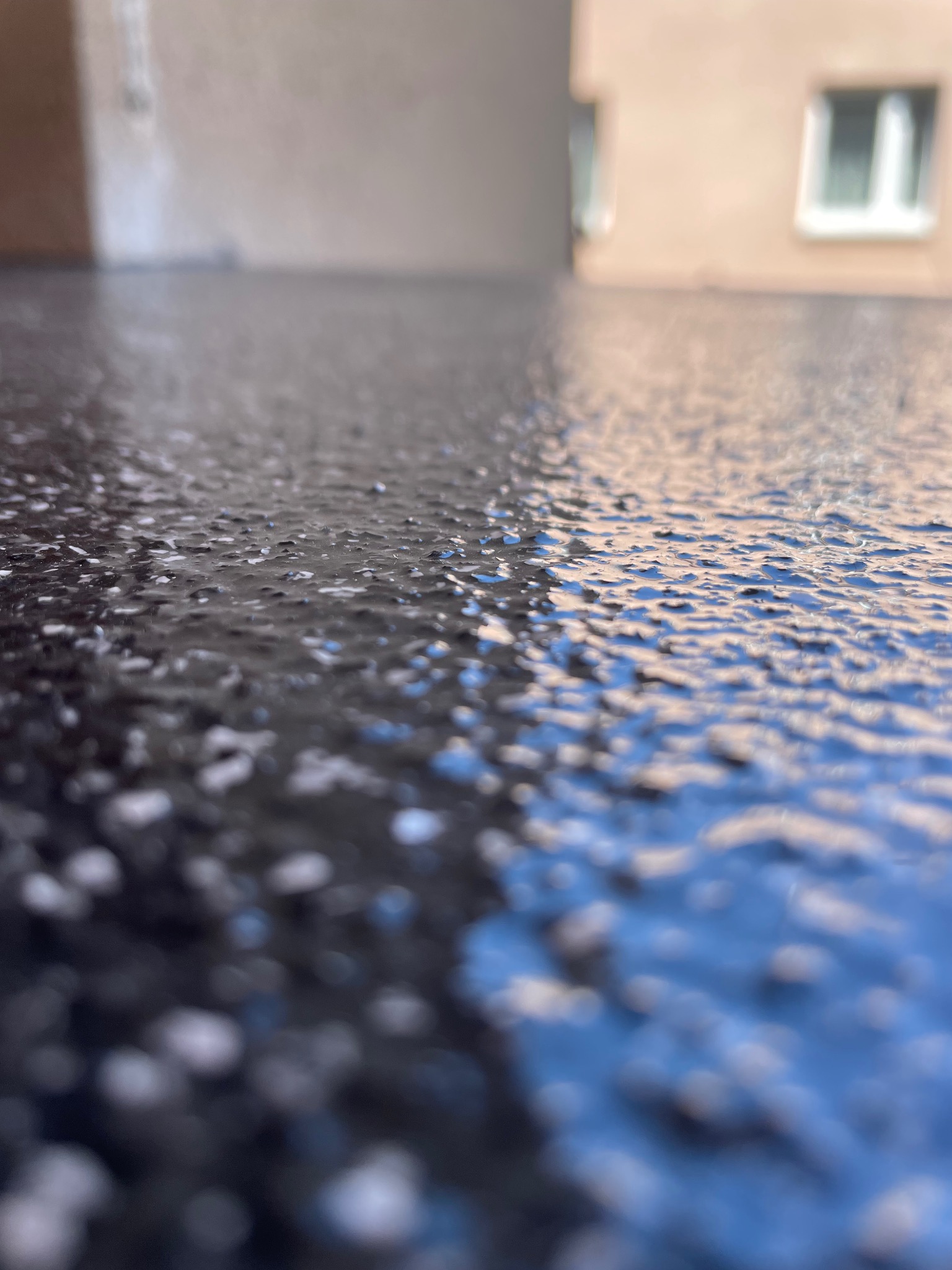 Błyszcząca, ciemna posadzka żywiczna z delikatną strukturą, sfotografowana pod kątem, z rozmytym tłem budynku z oknem.