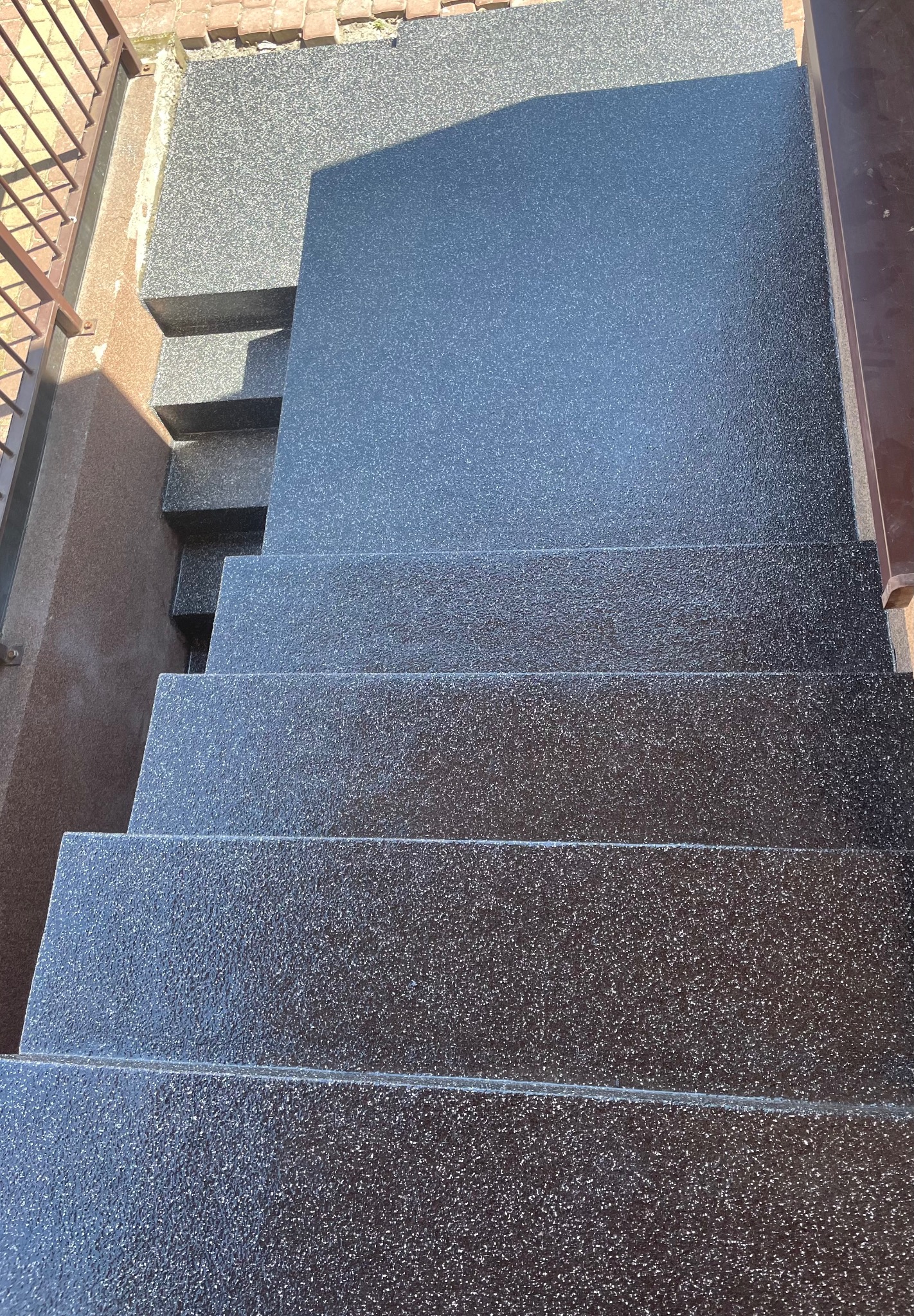 Schody zewnętrzne pokryte ciemną, błyszczącą powłoką z widoczną fakturą, z metalową barierką po prawej stronie i fragmentem ceglanego muru w tle.