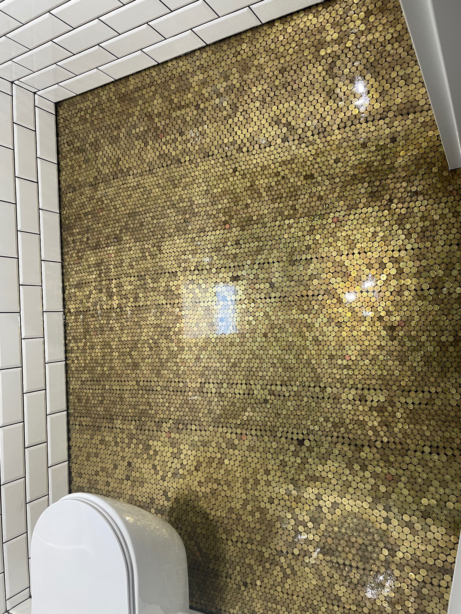 Podłoga w łazience wyłożona złotymi monetami zatopionymi w przezroczystej żywicy, widoczne białe płytki na ścianach i fragment toalety.