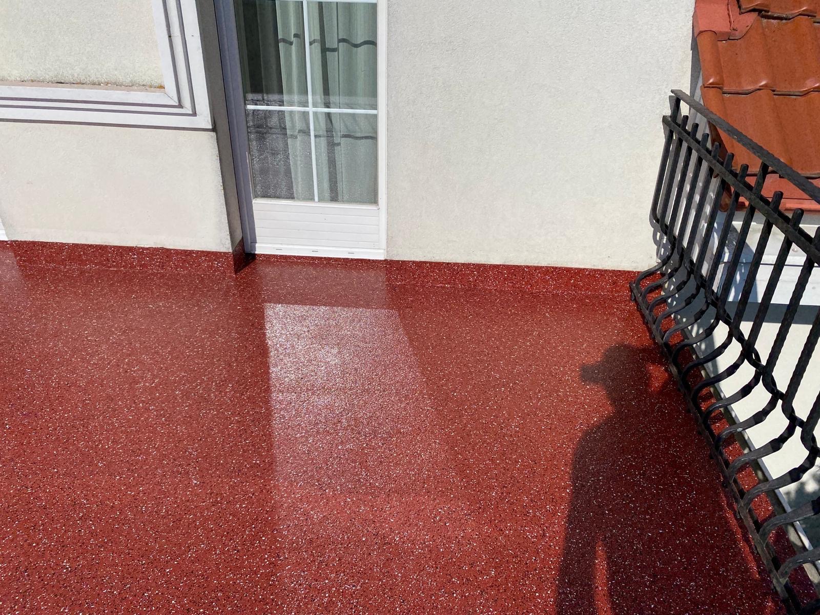 Balkon z czerwoną, błyszczącą posadzką z widoczną teksturą, cień osoby po prawej stronie, białe drzwi balkonowe i fragment metalowej balustrady.