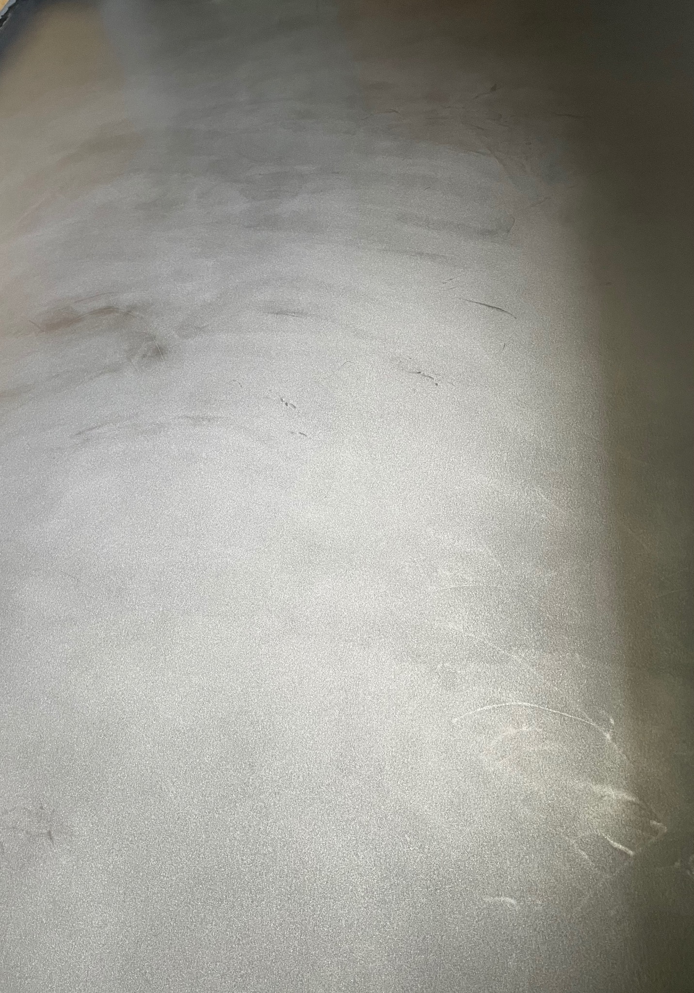 Jasnoszara, gładka powierzchnia z delikatnym połyskiem, przypominająca beton lub żywicę, z subtelnymi smugami i niedoskonałościami w fakturze, sfotografowana pod kątem.