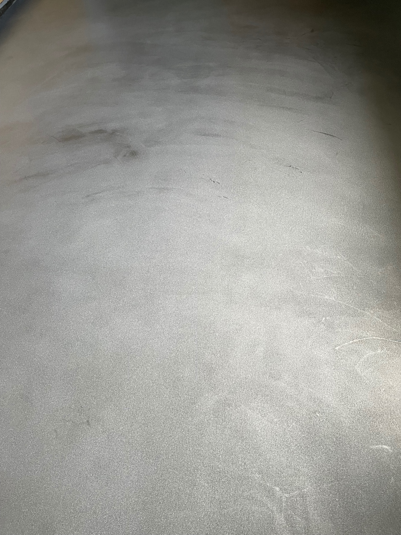 Szara, gładka powierzchnia podłogi z delikatnymi smugami i nierównomiernym rozkładem światła, przypominająca efekt polerowanego betonu.