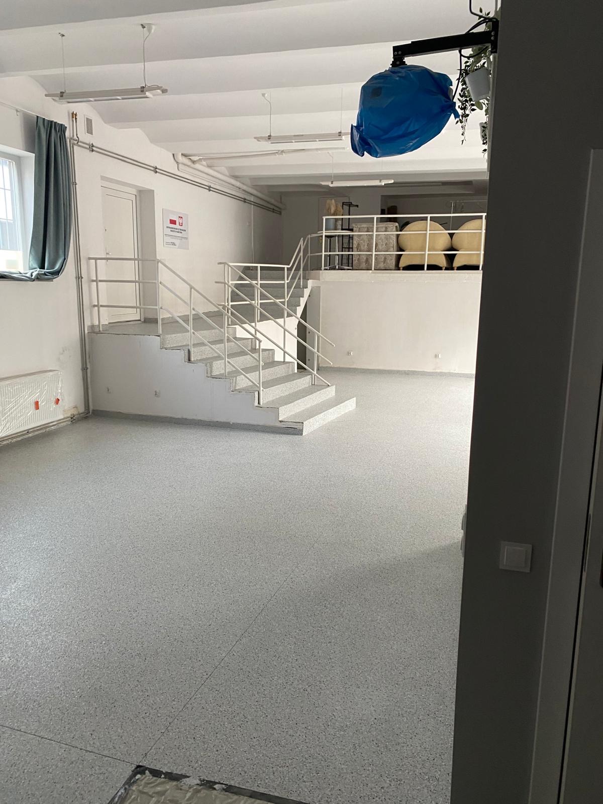 Jasnoszara posadzka z drobnymi plamkami w przestronnym wnętrzu z białymi schodami i balustradami. Widoczne okno z zasłoną i elementy instalacji pod sufitem.