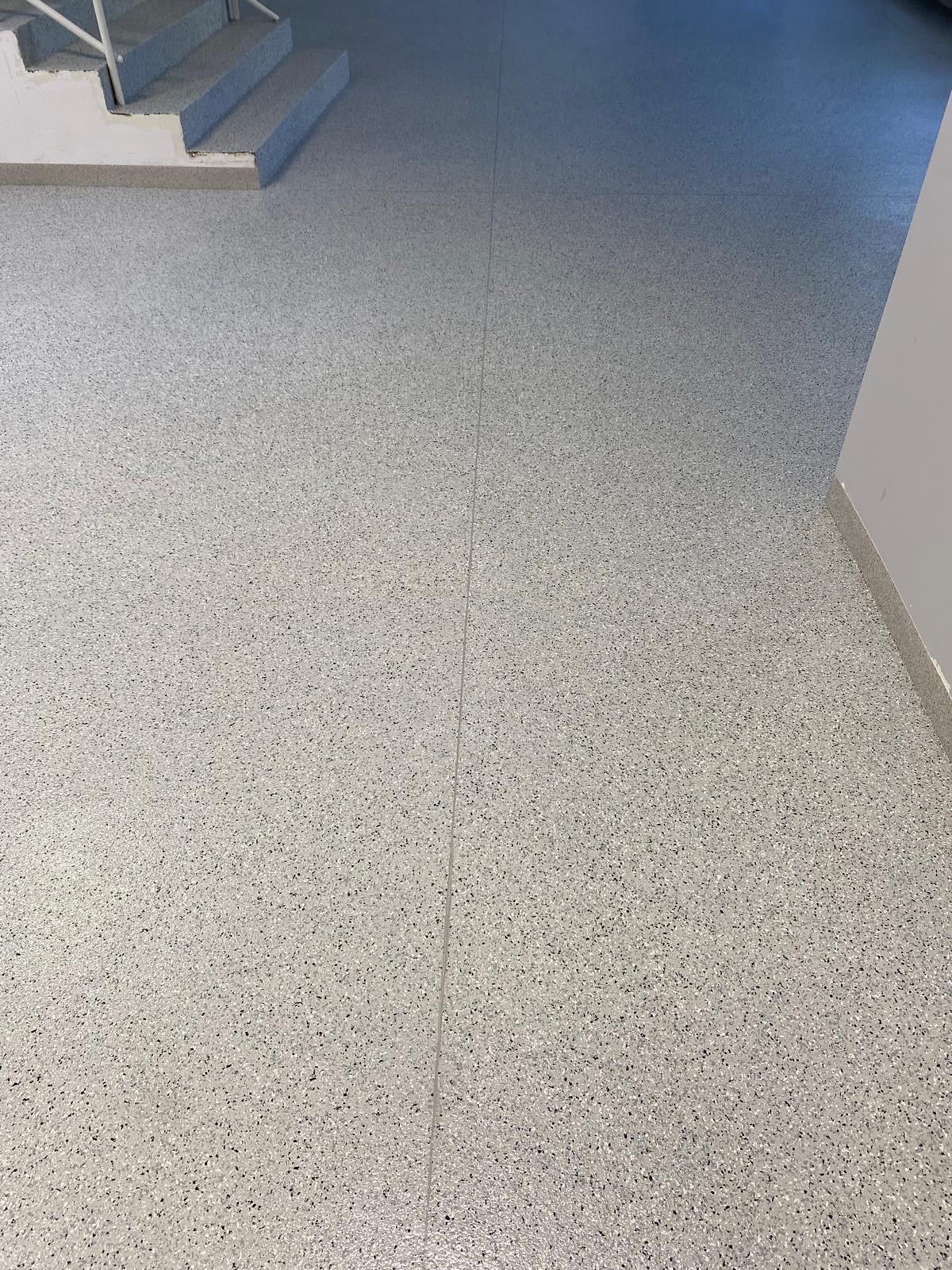 Jasnoszara posadzka z drobnymi, ciemnymi plamkami, zbliżenie na fragment schodów i ściany. Gładka powierzchnia, widoczne łączenia płyt. Stonowana kolorystyka.