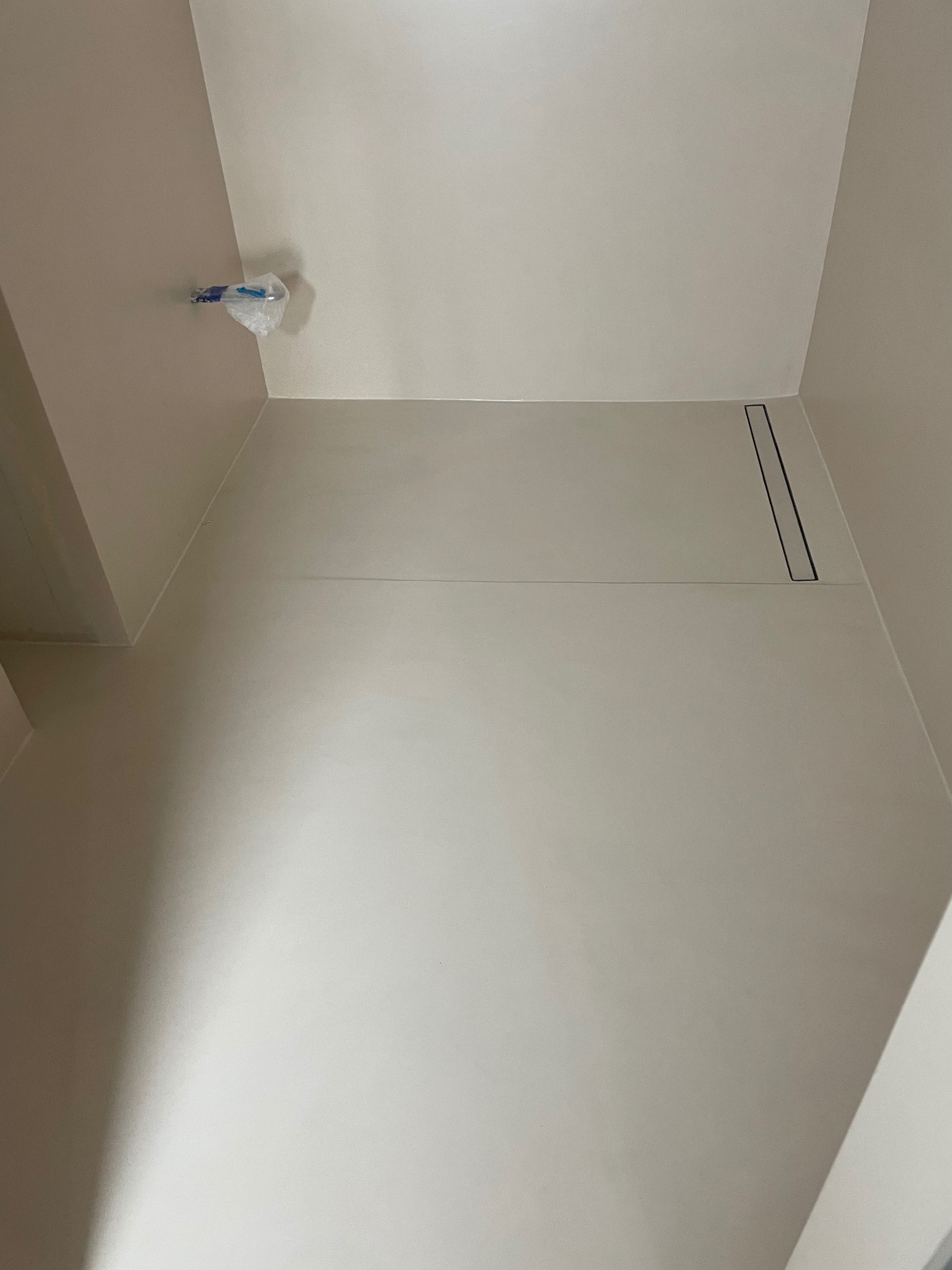 Minimalistyczna łazienka w odcieniach beżu z odpływem liniowym w podłodze. Widoczne zabezpieczenie instalacji na ścianie. Gładkie powierzchnie ścian i podłogi.