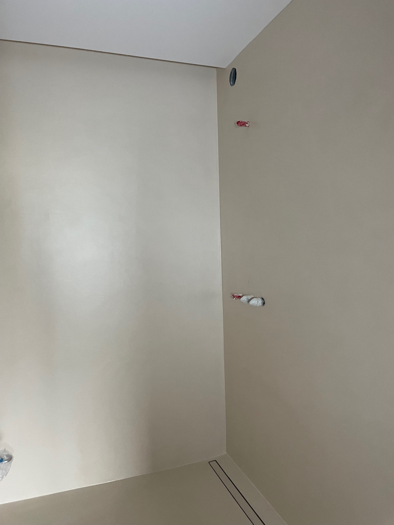 Beżowe ściany i podłoga w narożniku pomieszczenia z widocznymi elementami instalacji. Minimalistyczny design, gładkie powierzchnie, nowoczesne wykończenie.