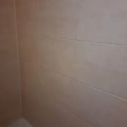 Beżowe płytki ceramiczne ułożone na ścianie w łazience, widoczny fragment wanny, szare fugi.