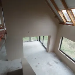 Wysoki pokój z tynkami maszynowymi w odcieniach beżu, widok z góry na podłogę z widocznymi śladami po pracach, okna dachowe i duże okna panoramiczne z widokiem na zewnątrz, rury instalacyjne...