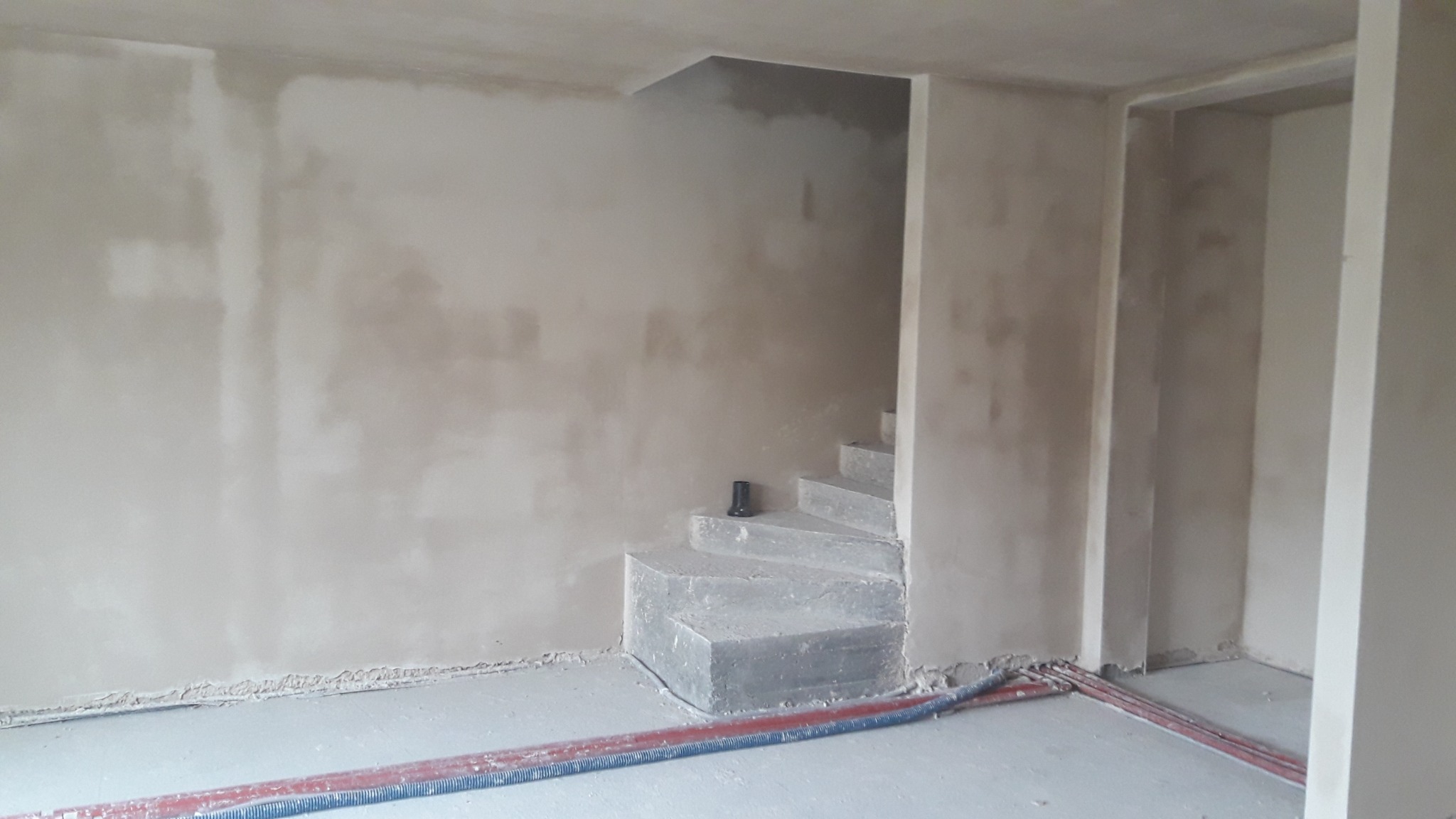 Świeżo położone tynki na ścianach wewnętrznych pomieszczenia ze schodami, widoczne przewody na podłodze i czarny element na schodach.