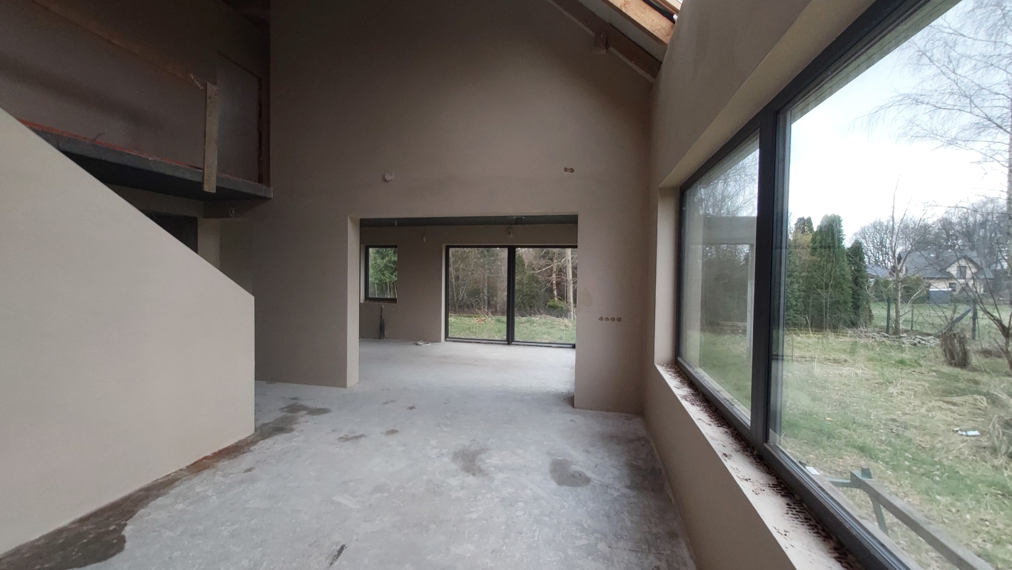 Surowe wnętrze domu w stanie deweloperskim po nałożeniu tynków maszynowych, z dużymi oknami z widokiem na zewnątrz i surową betonową podłogą.