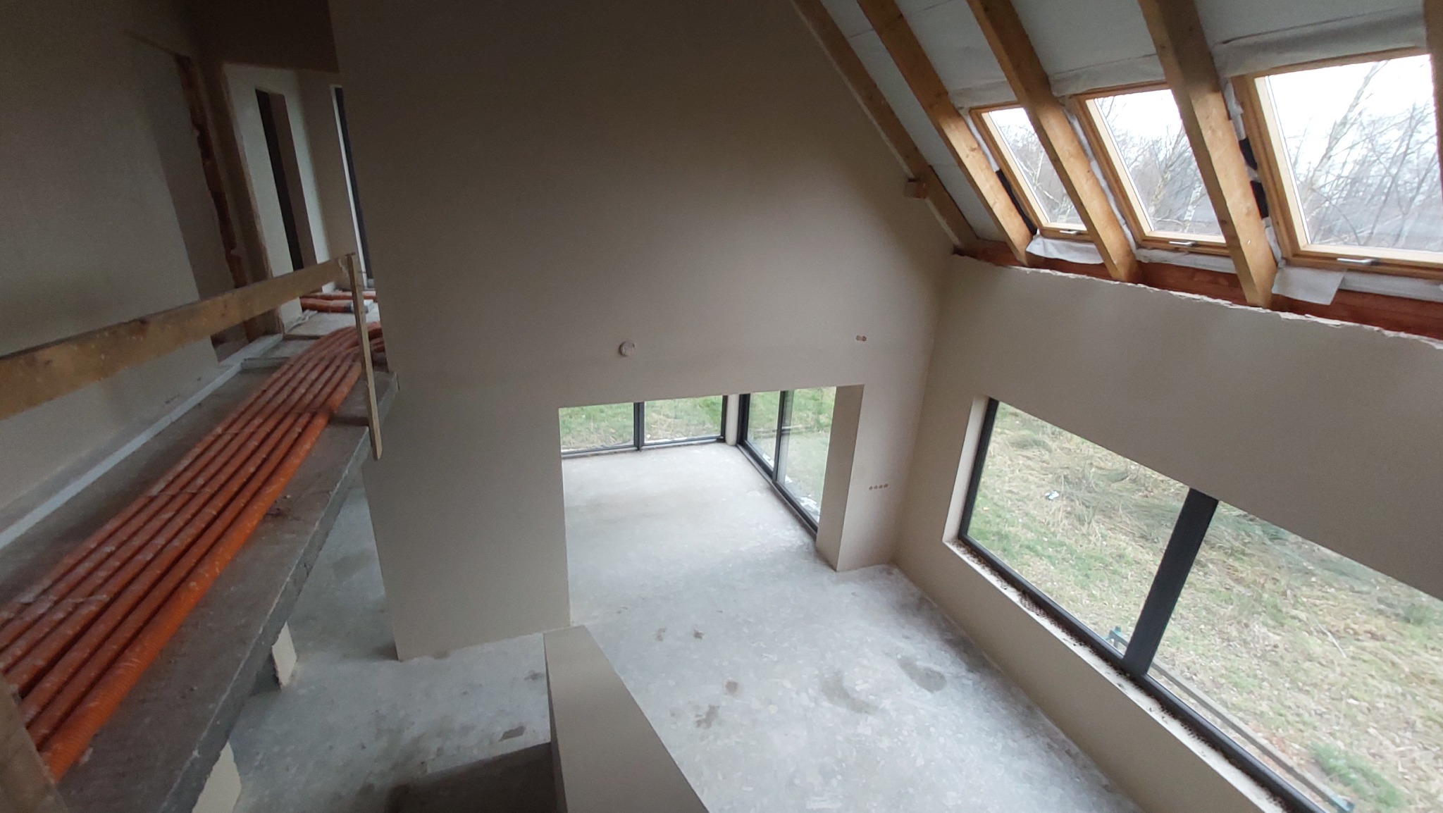 Wysoki pokój z tynkami maszynowymi w odcieniach beżu, widok z góry na podłogę z widocznymi śladami po pracach, okna dachowe i duże okna panoramiczne z widokiem na zewnątrz, rury instalacyjne...