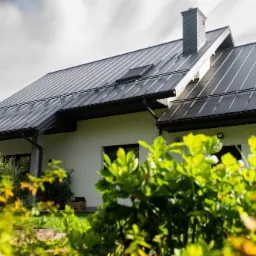 Nowoczesny dom jednorodzinny w Dębicy z ciemnoszarą blachą na dachu, rynnami i kominem, widoczny z perspektywy ogrodu z bujną zielenią.