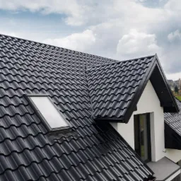 Czarny dach z dachówki modułowej z oknem dachowym, widok z góry na nowy dom z białymi ścianami i czarnymi ramami okiennymi, w tle zielone pola i pochmurne niebo.