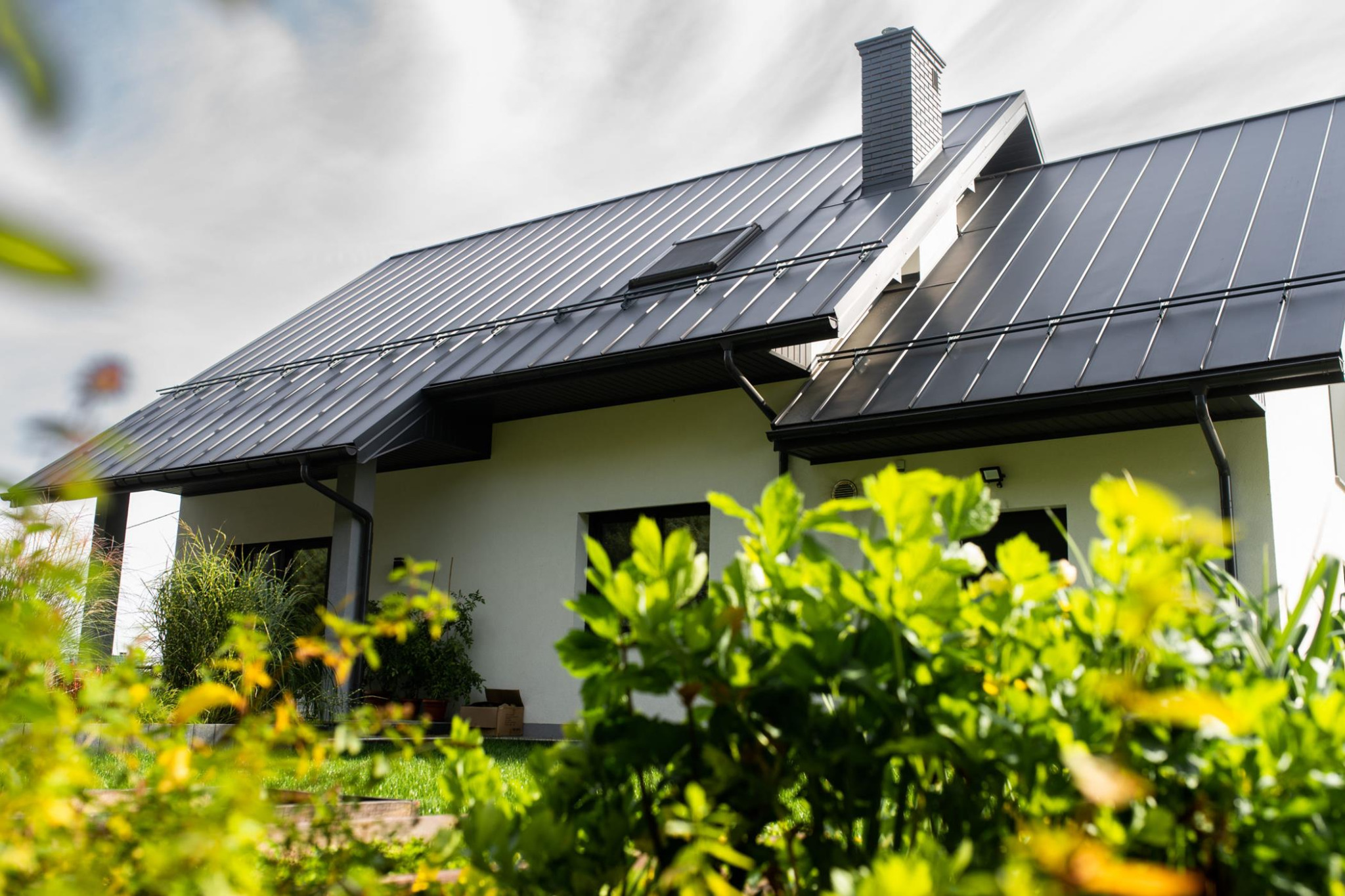 Nowoczesny dom jednorodzinny w Dębicy z ciemnoszarą blachą na dachu, rynnami i kominem, widoczny z perspektywy ogrodu z bujną zielenią.