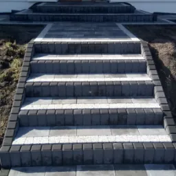 Schody wejściowe do domu wyłożone kostką brukową w odcieniach szarości i bieli, z widoczną fakturą materiału i świeżo ułożonym gruntem po bokach.