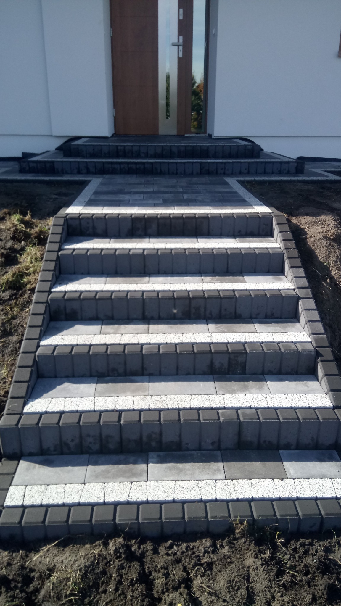 Schody wejściowe do domu wyłożone kostką brukową w odcieniach szarości i bieli, z widoczną fakturą materiału i świeżo ułożonym gruntem po bokach.