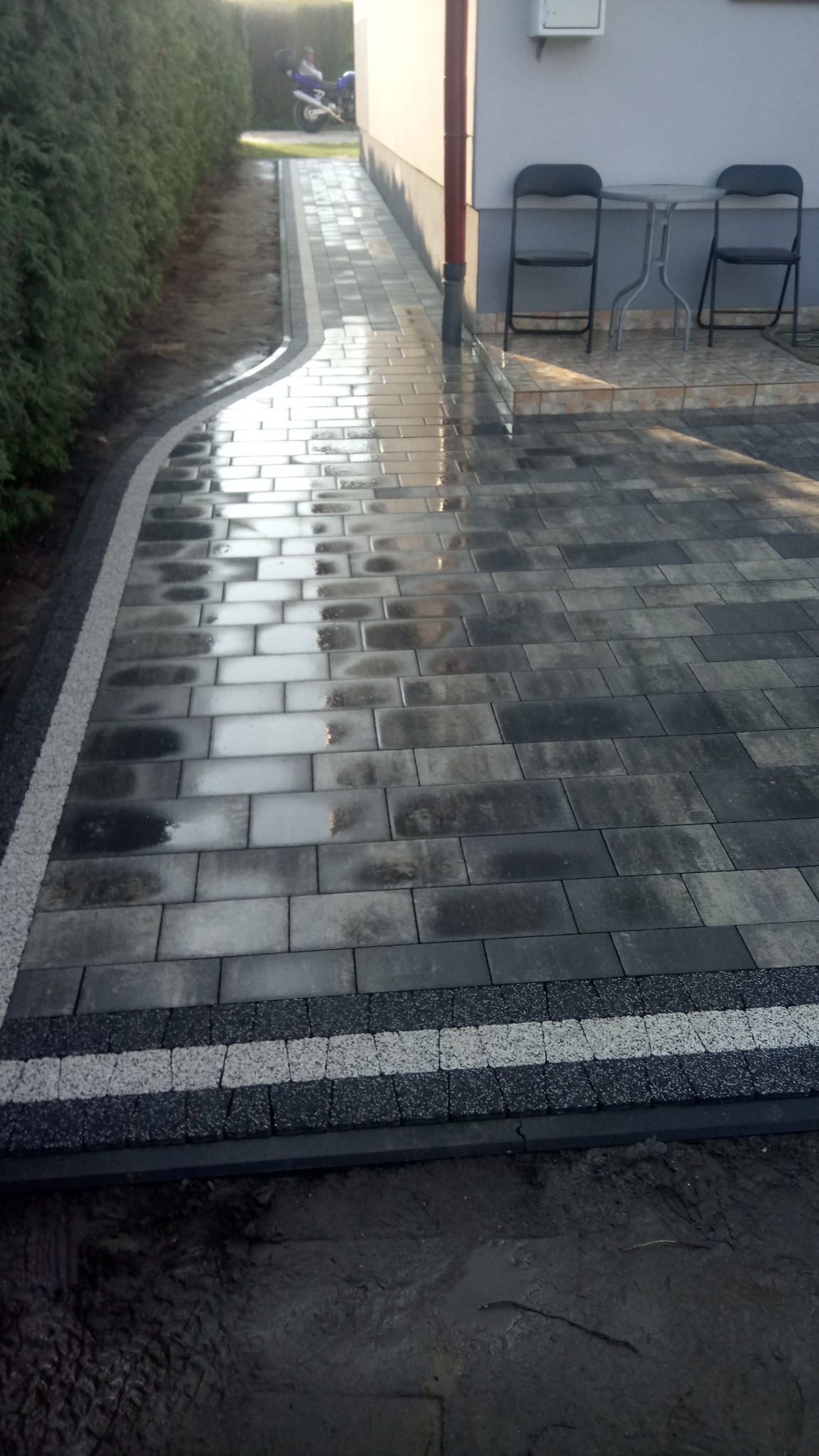 Świeżo ułożona, mokra, szara kostka brukowa z dekoracyjnym obramowaniem z jasnego i ciemnego grysu, prowadząca wzdłuż budynku z szarymi ścianami i zielonym żywopłotem, z motocyklem w tle.