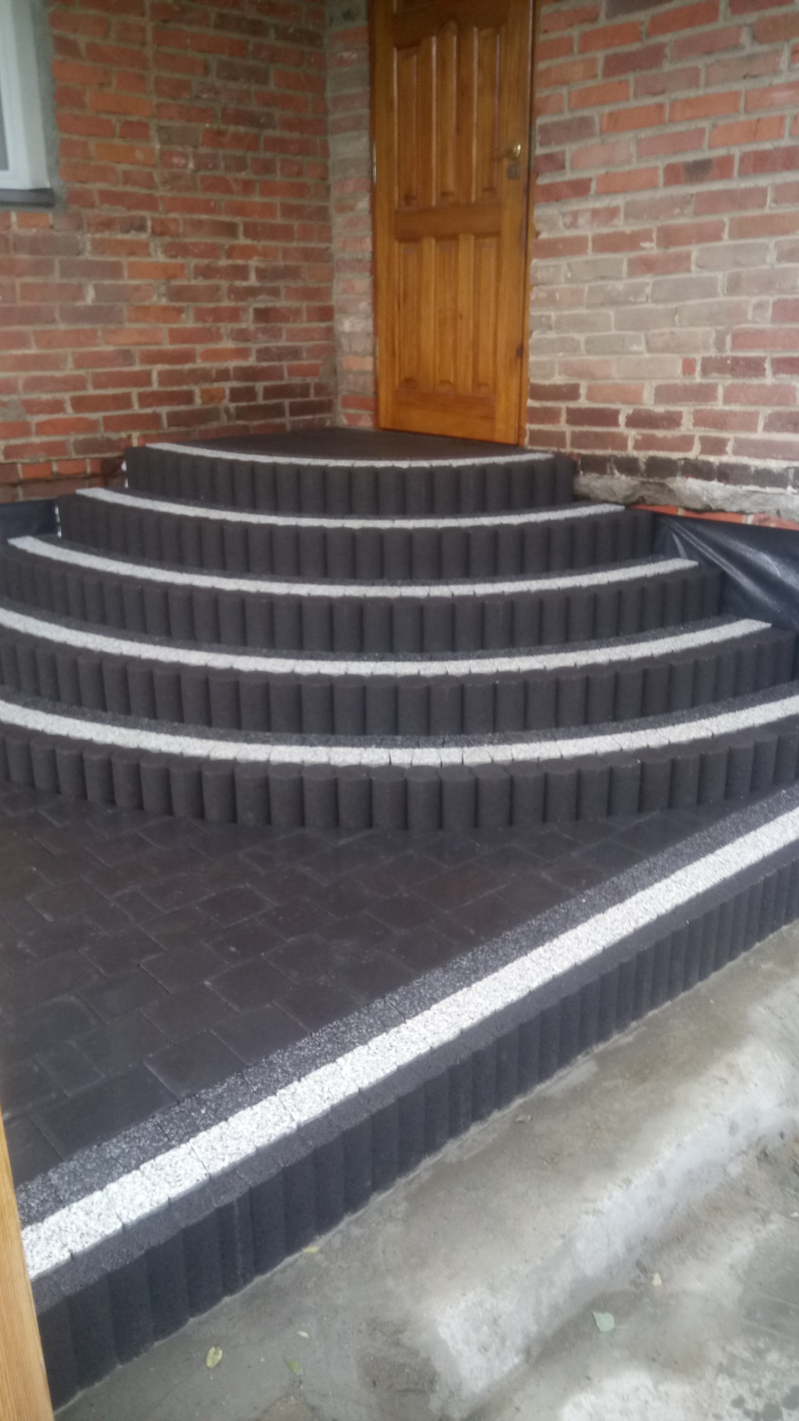 Schody wejściowe wyłożone ciemną kostką brukową z jasnymi paskami, prowadzące do drewnianych drzwi w budynku z czerwonej cegły.