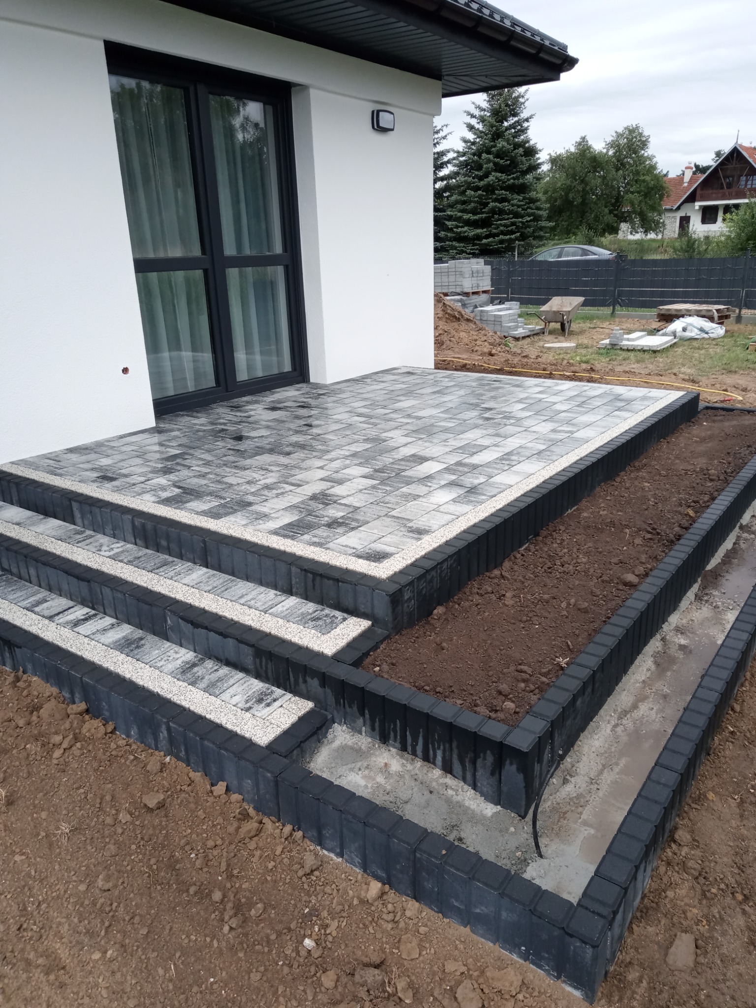 Nowoczesny taras z kostki brukowej w odcieniach szarości, z trzema stopniami i obramowaniem z czarnej kostki. Przy tarasie widoczna rabata kwiatowa z czarnym obrzeżem, wypełniona ziemią. W tle...