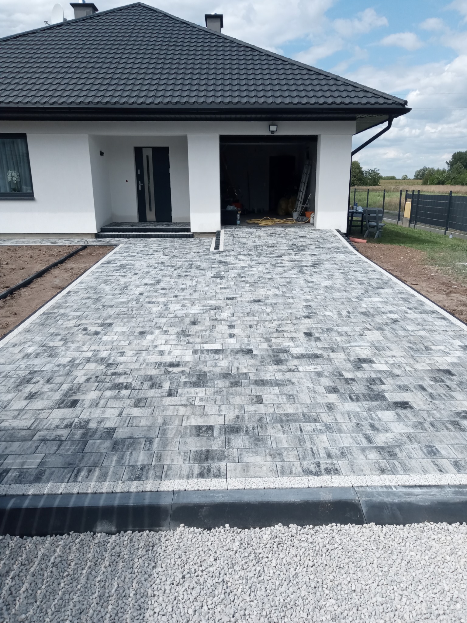 Ułożona szara kostka brukowa na podjeździe do nowoczesnego domu z czarnym dachem i białymi ścianami, widok z góry na gotową realizację.