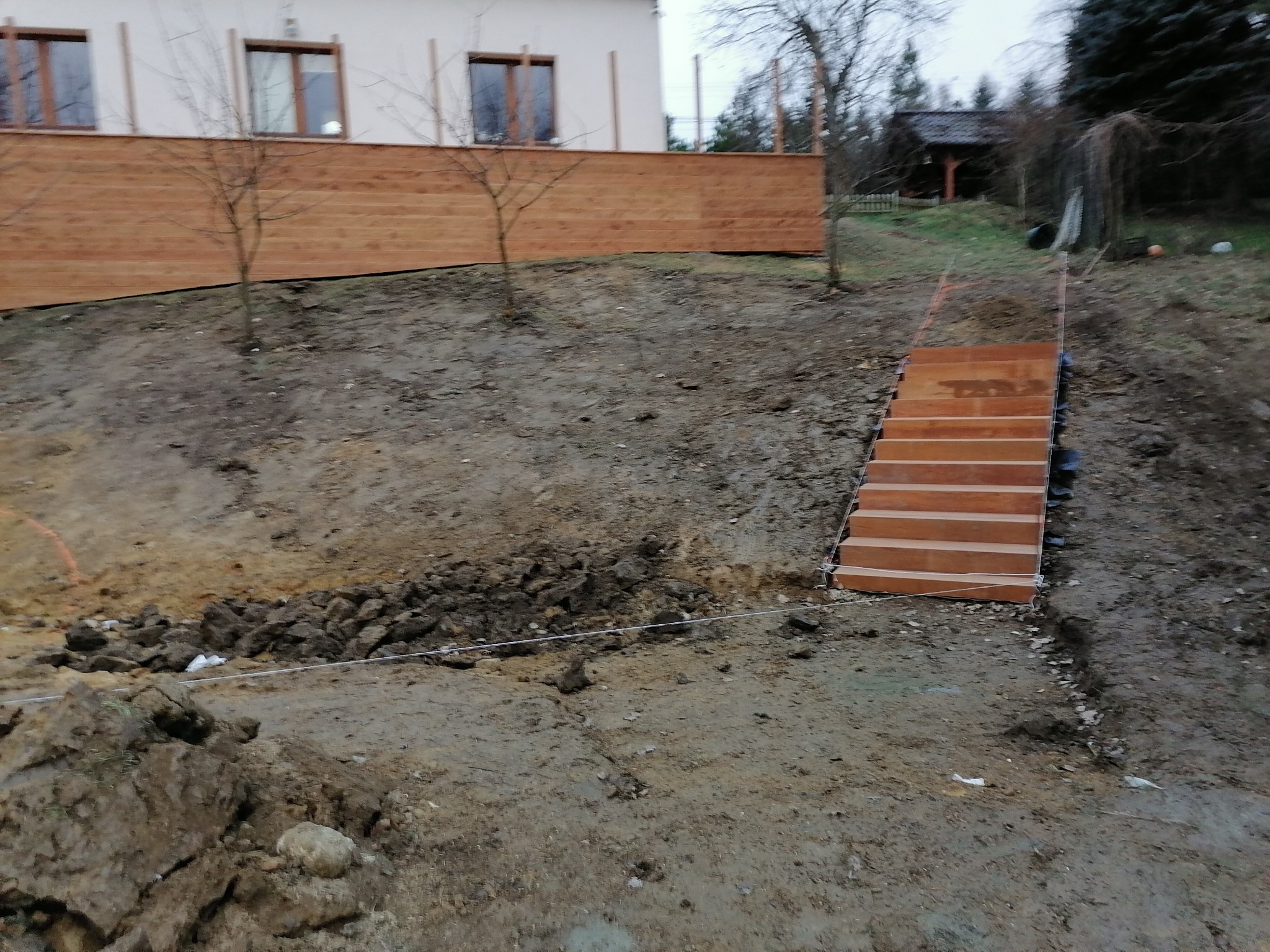 Budowa drewnianych schodów zewnętrznych w ogrodzie, widok na częściowo wykopany teren, drewniany płot i dom w tle.