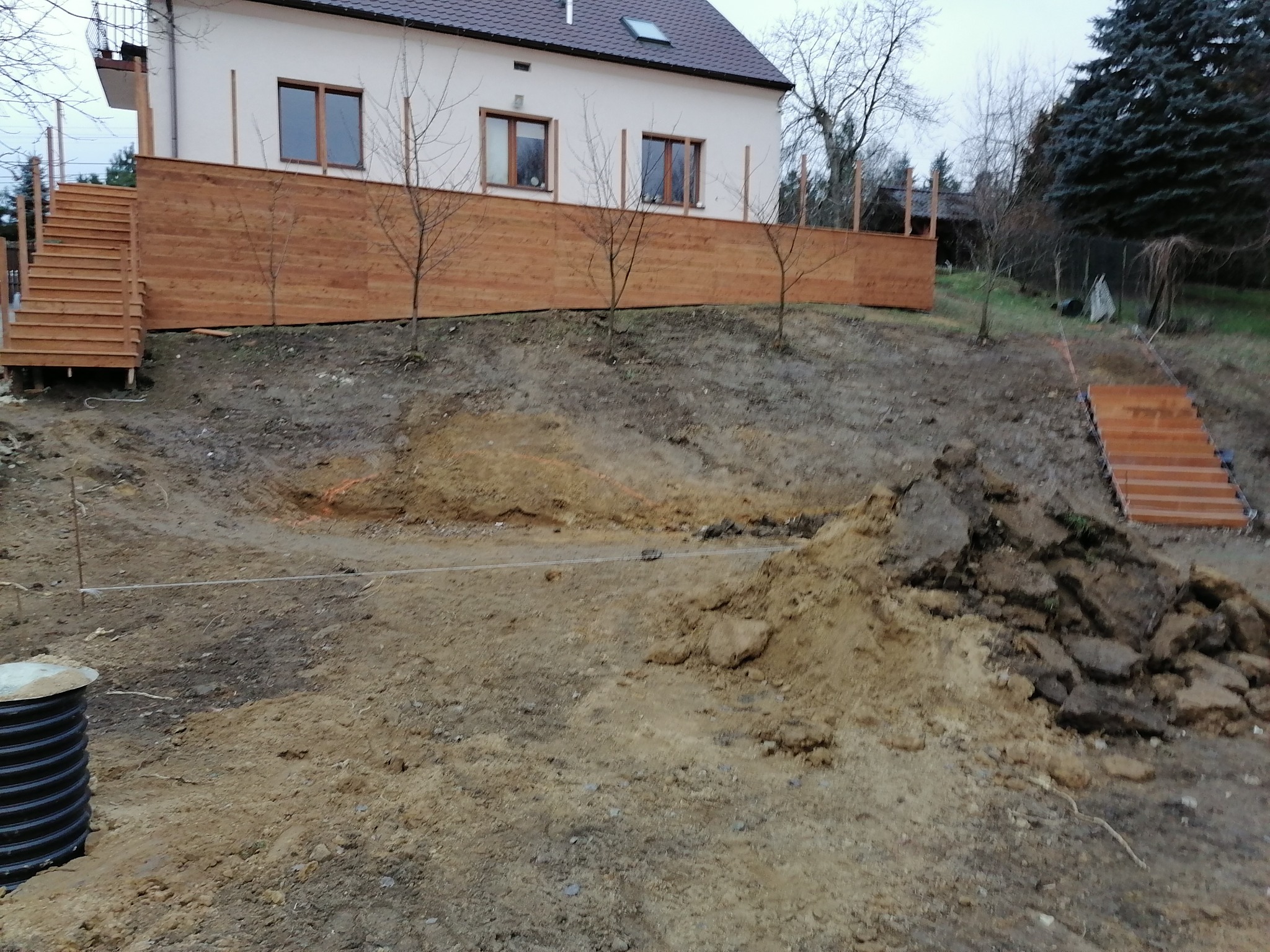 Przygotowanie terenu pod budowę tarasu: wykopane fundamenty, widoczne deski szalunkowe, drewniane schody prowadzące na górę, dom w tle.
