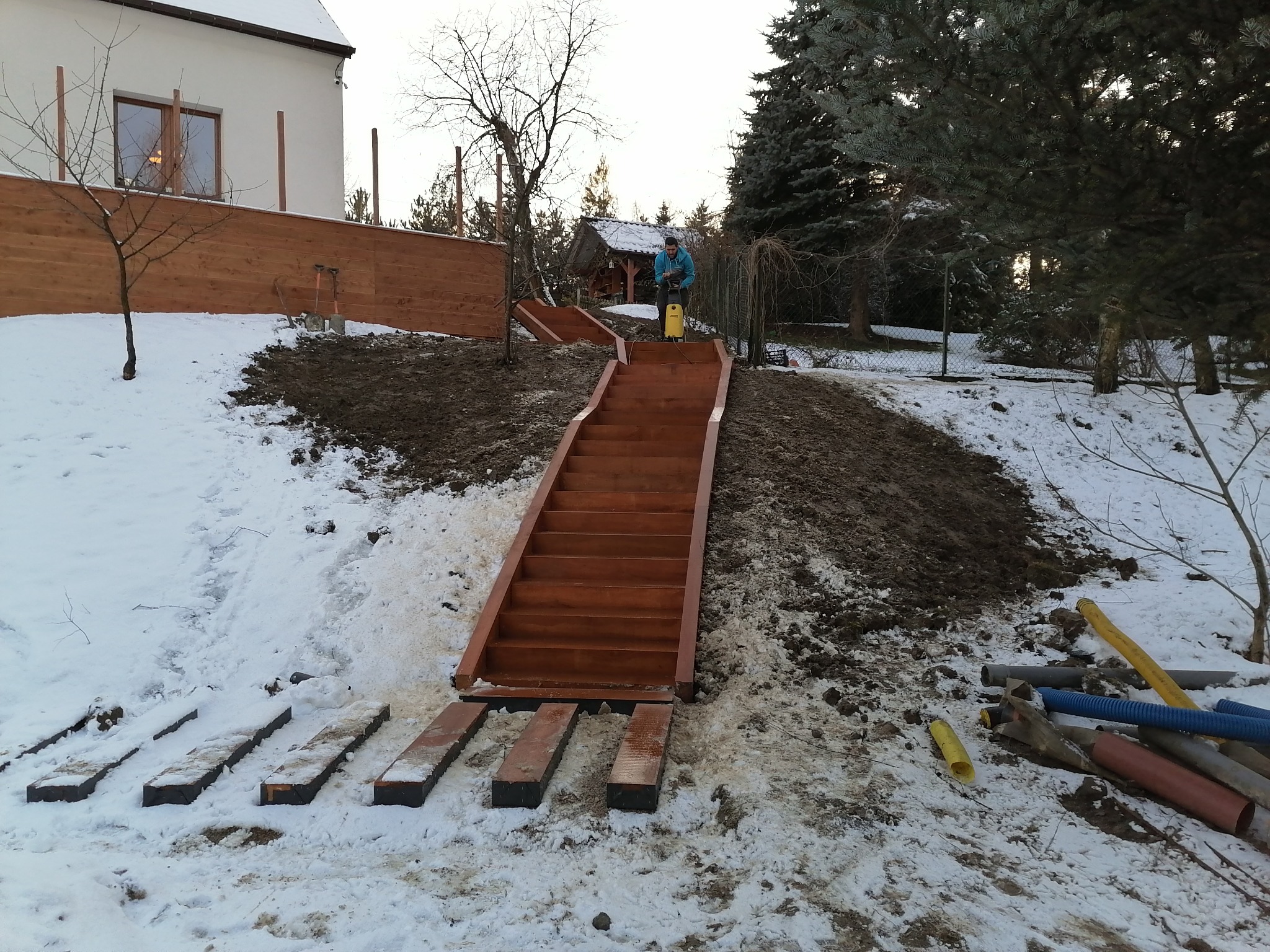 Montaż drewnianych schodów zewnętrznych na zboczu w zimowej scenerii; pracownik z myjką ciśnieniową na górze schodów.