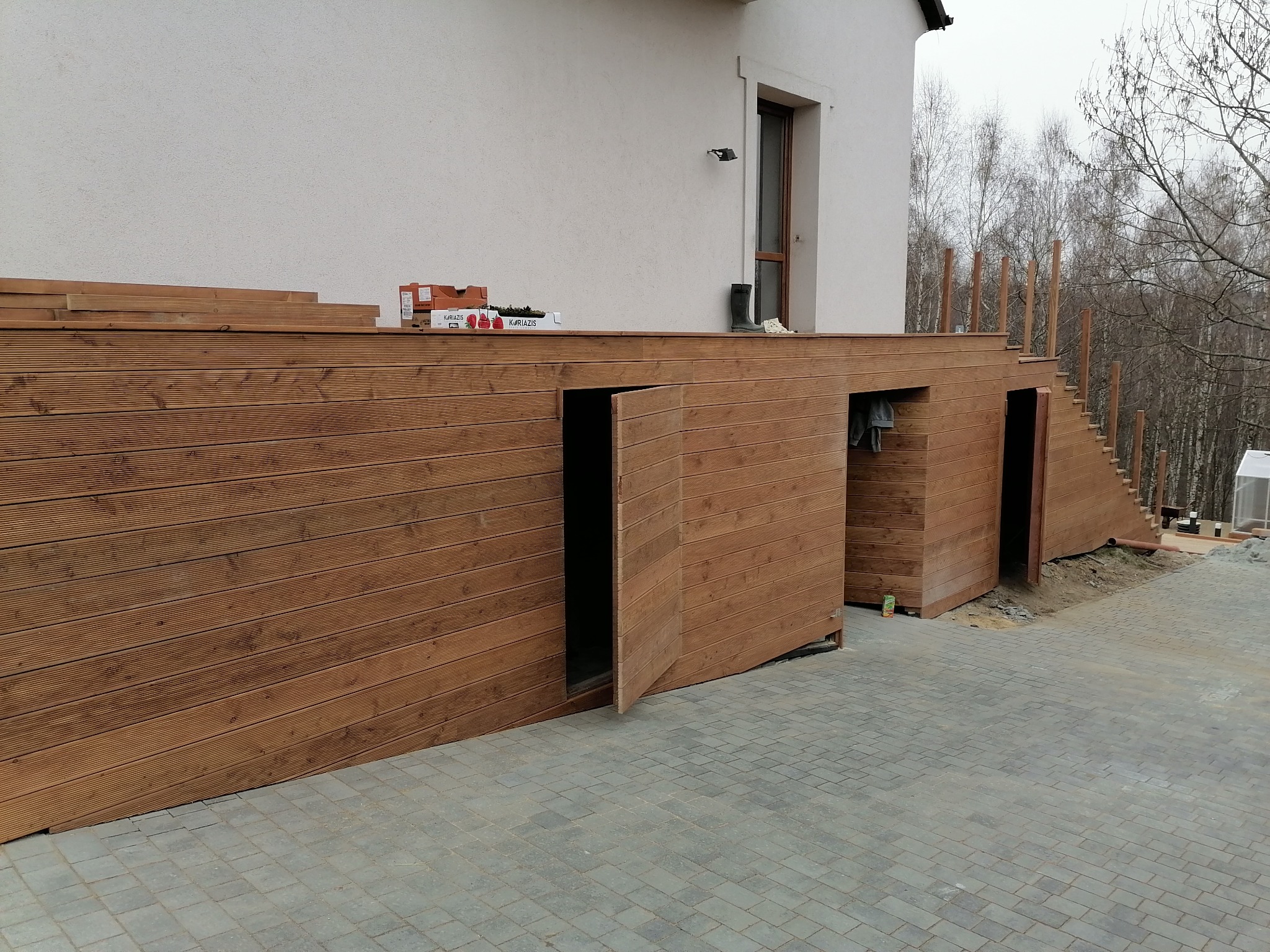 Drewniany taras z ukrytymi schowkami i schodami, widok z poziomu kostki brukowej. Na tarasie skrzynki z owocami i guma kalosza przy oknie.