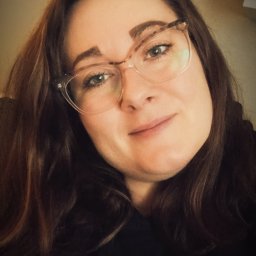 JUSTYNA KOSYK - Portret kobiety w okularach z brązowymi włosami i symbolem pacyfistycznym na czarnej bluzce, uśmiechająca się do kamery.