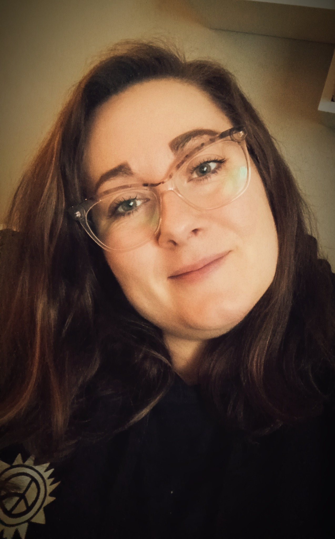 Portret kobiety w okularach z brązowymi włosami i symbolem pacyfistycznym na czarnej bluzce, uśmiechająca się do kamery.