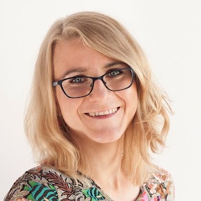 Uśmiechnięta blondynka w okularach, z kolczykiem w nosie, w kolorowej bluzce na białym tle. Portret kobiety.