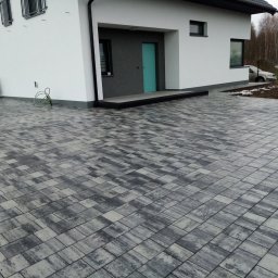 BrukPol - Nowoczesny dom jednorodzinny z podjazdem wyłożonym szarą kostką brukową w geometryczny wzór. W tle widoczna osoba i sprzęt klimatyzacyjny.