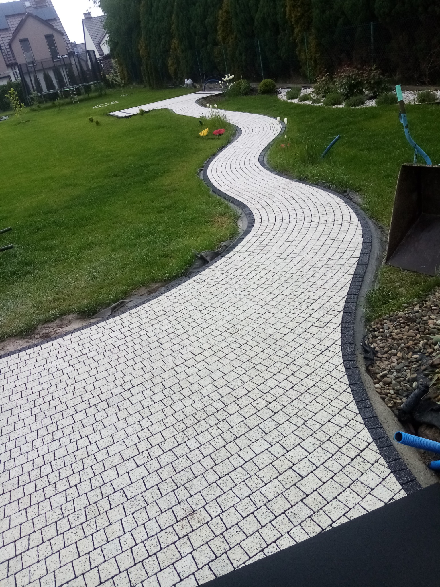 Kremowa ścieżka z kostki brukowej z ciemnoszarym obramowaniem, wijąca się przez zielony trawnik w ogrodzie, z widocznym fragmentem łyżki koparki i niebieskim wężem, wskazującym na trwające prace...