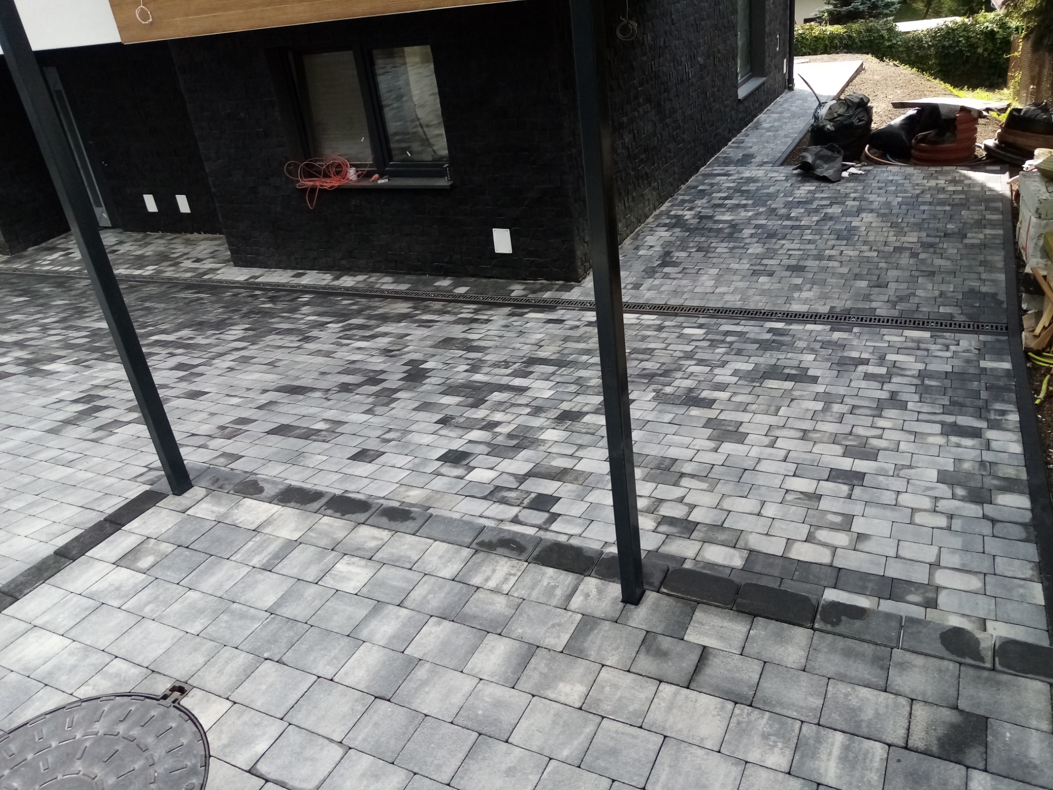 Ułożona szaro-czarna kostka brukowa na podjeździe z odpływem liniowym, widok z góry, w tle czarny budynek z oknem i stertą materiałów budowlanych.