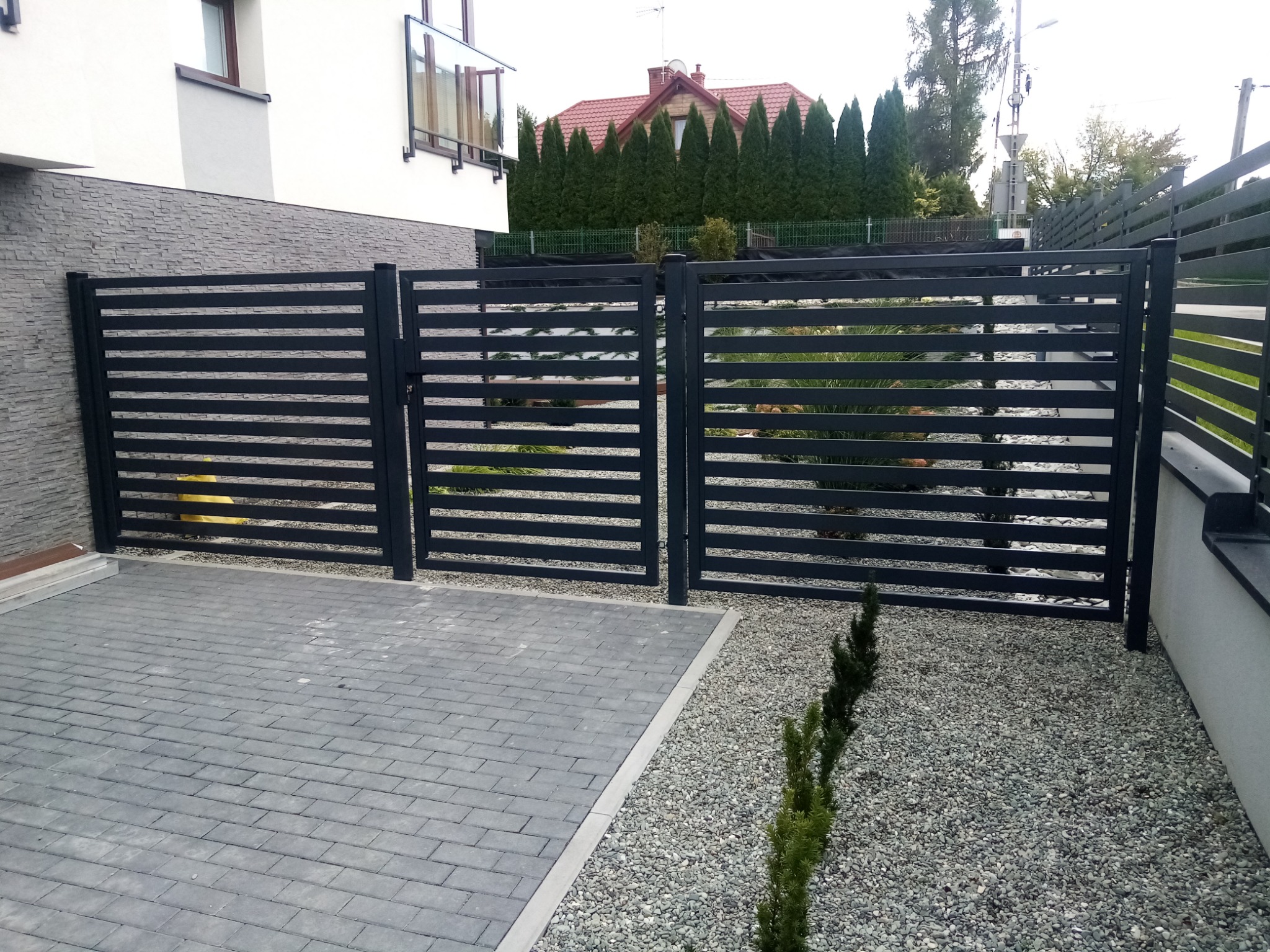 Nowoczesne, poziome przęsła metalowe tworzące ogrodzenie i furtkę, widoczne z poziomu brukowanego podjazdu, z dekoracyjnym obsypaniem kamieniami i młodymi iglakami.