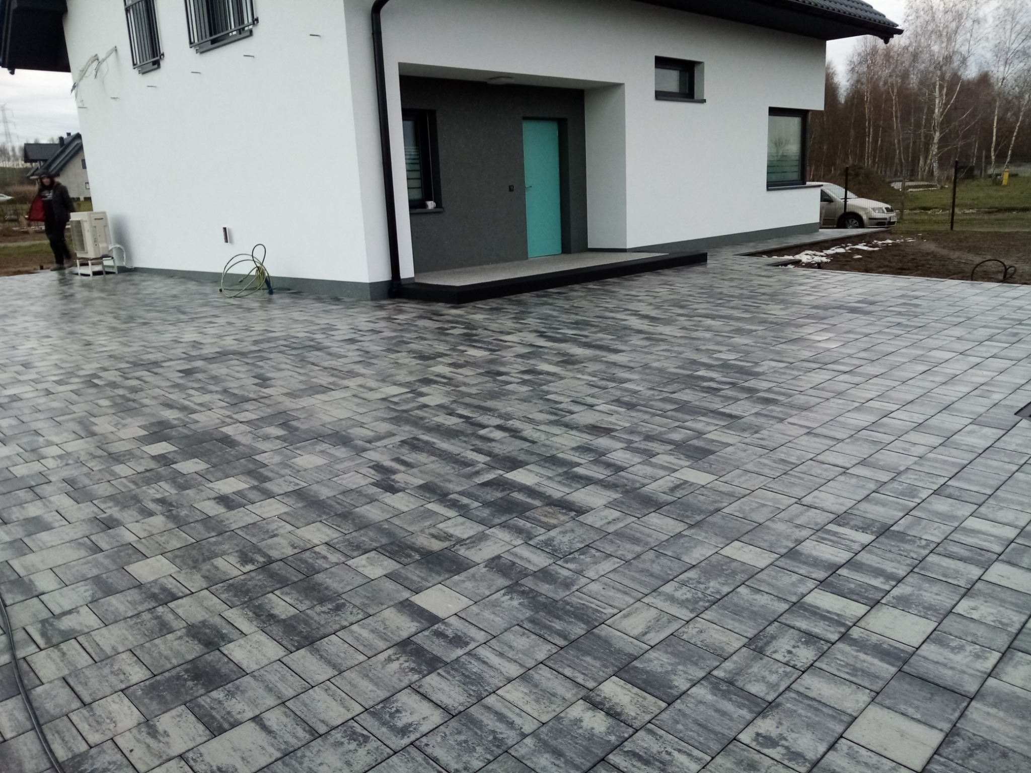 Nowoczesny dom jednorodzinny z podjazdem wyłożonym szarą kostką brukową w geometryczny wzór. W tle widoczna osoba i sprzęt klimatyzacyjny.