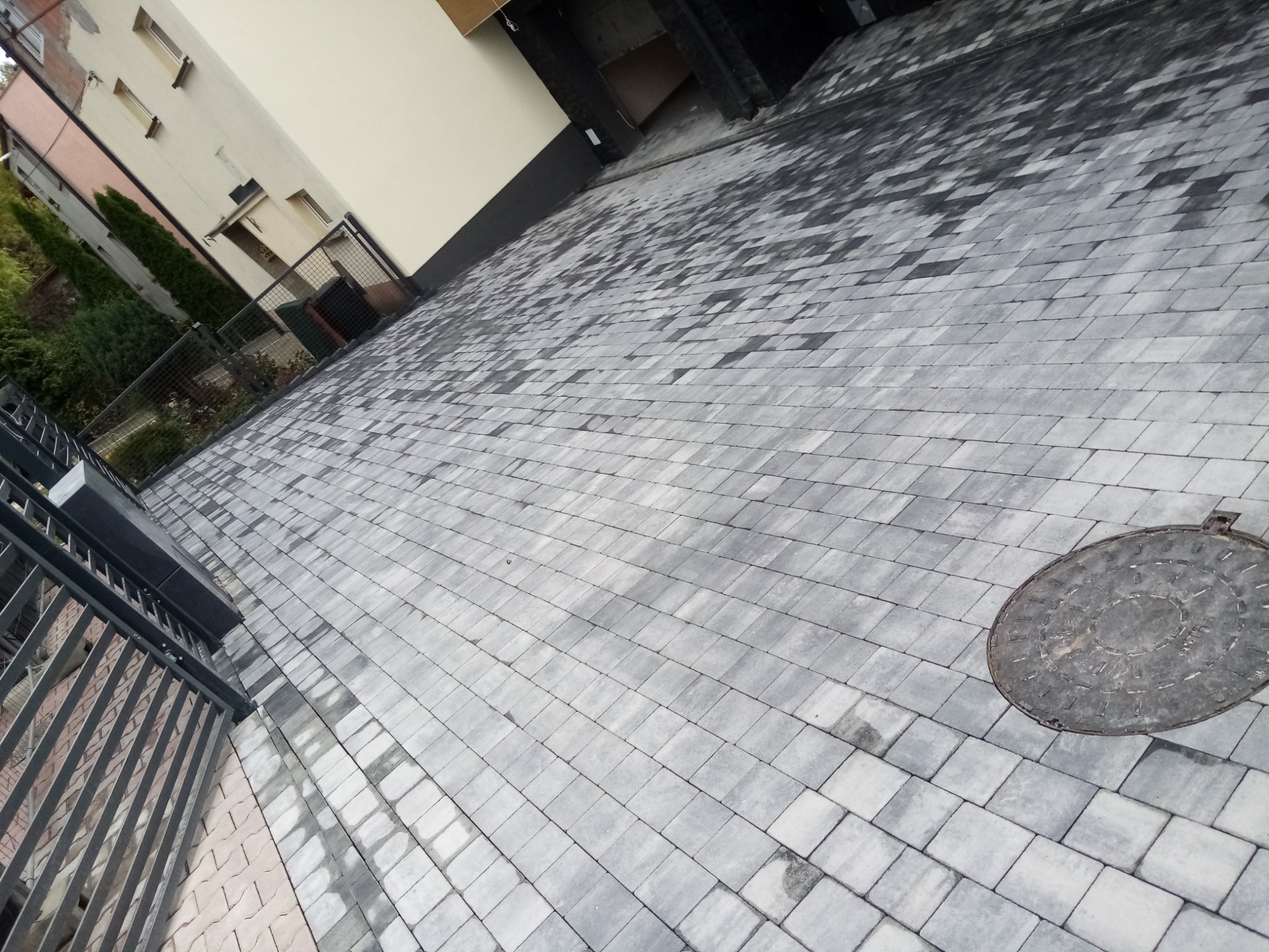 Nowa, szara kostka brukowa ułożona na podjeździe, widoczna studzienka kanalizacyjna i fragment metalowego ogrodzenia.