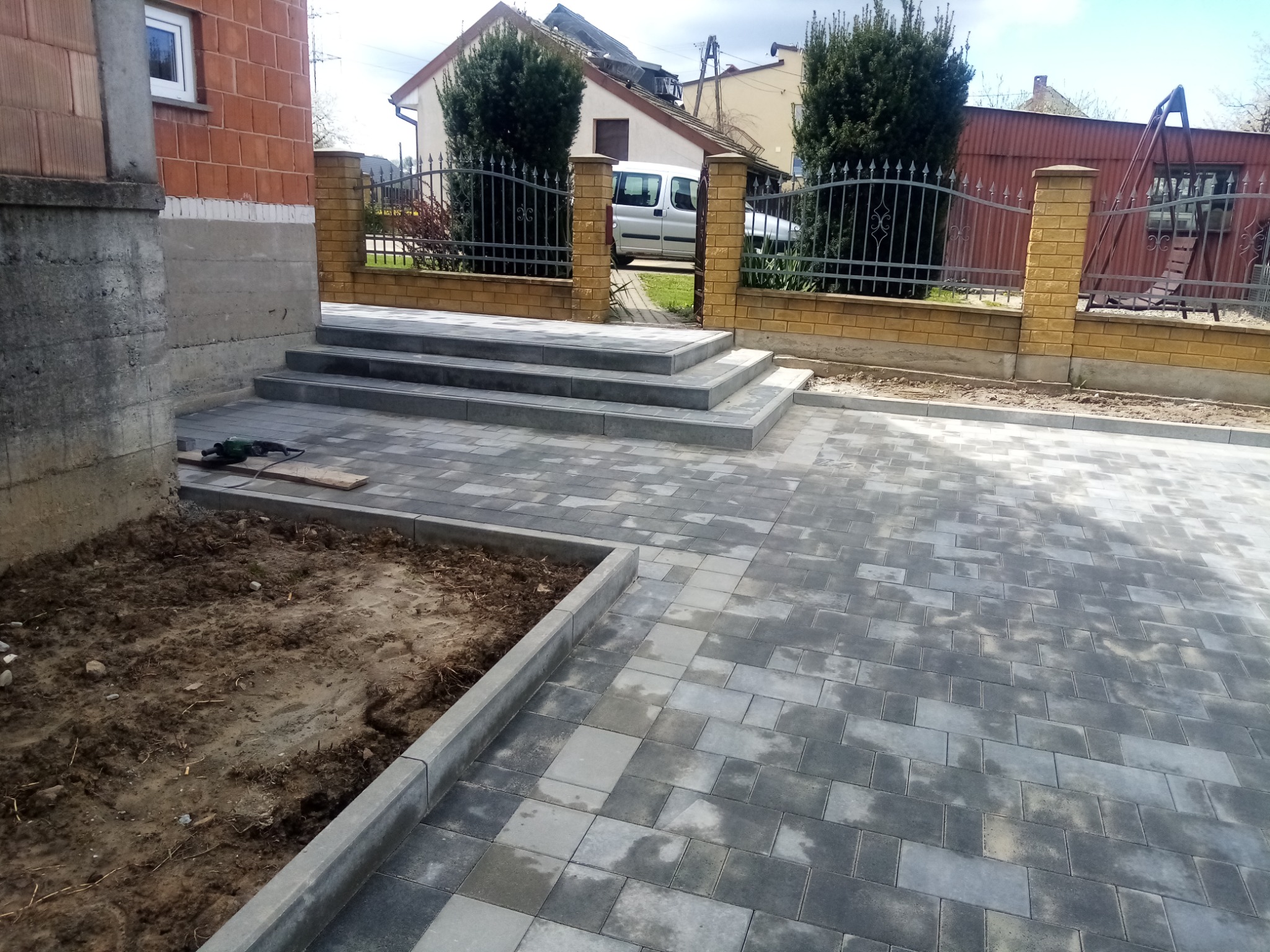Świeżo ułożona, szara kostka brukowa na podjeździe i schodach wejściowych do domu, z widocznym fragmentem niezabudowanej ziemi i narzędziem elektrycznym w rogu.