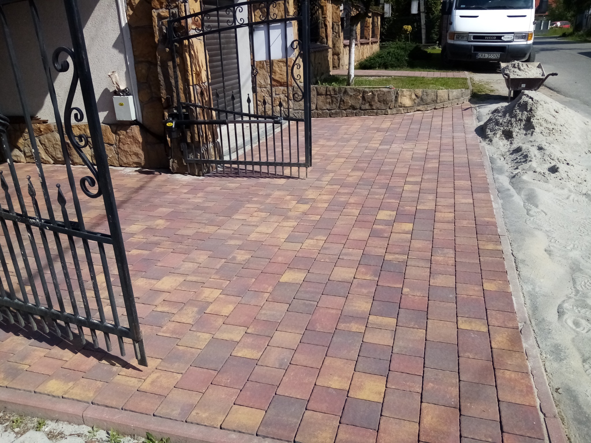 Nowo ułożona, wielokolorowa kostka brukowa prowadząca od otwartej, ozdobnej bramy wjazdowej, z widoczną stertą piasku i taczką w tle oraz fragmentem zaparkowanego białego dostawczaka.