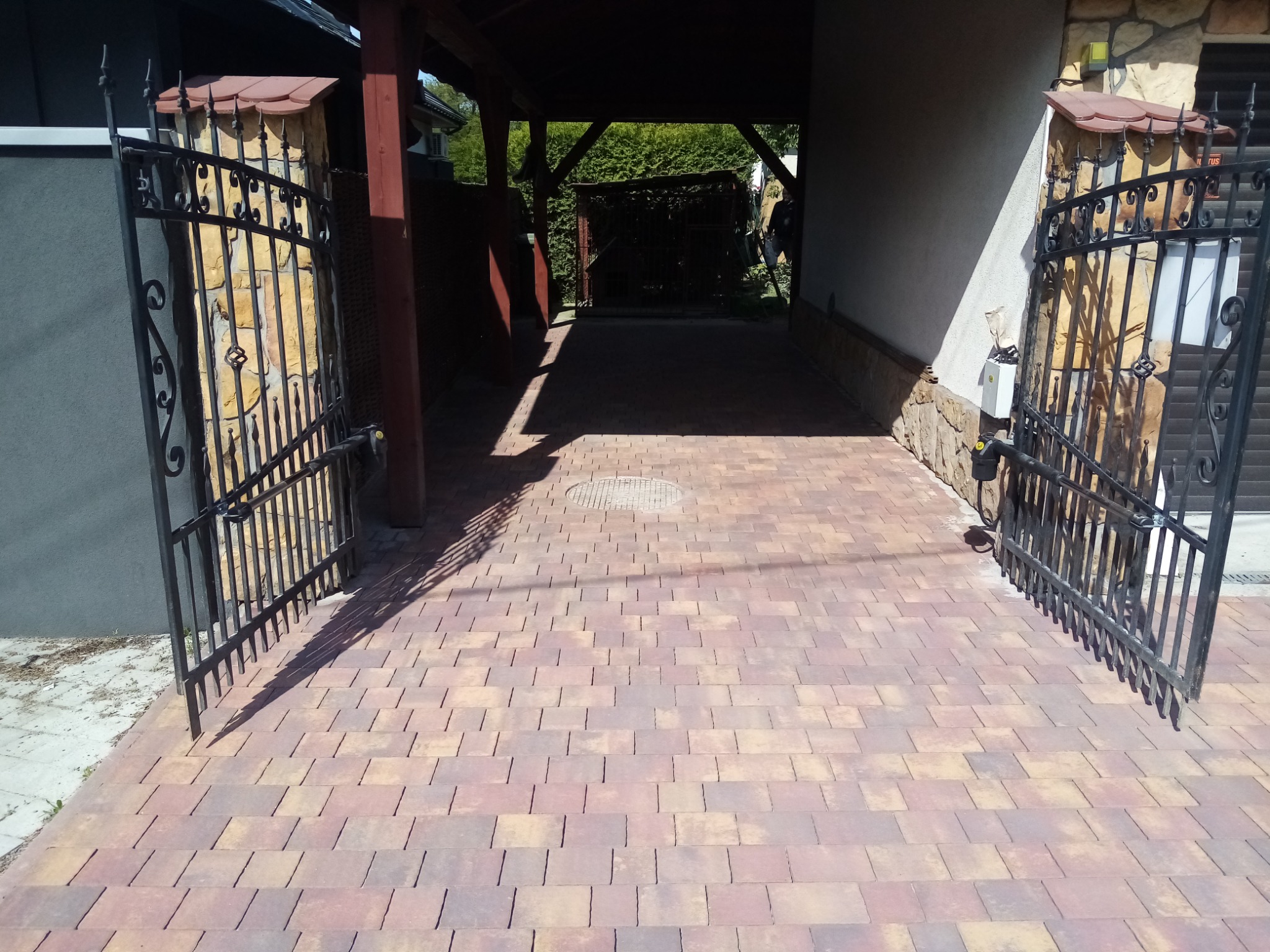 Otwarta, czarna, metalowa brama wjazdowa z dekoracyjnymi elementami, osadzona na kamiennych słupkach z daszkami, prowadząca na podjazd wyłożony kostką w odcieniach czerwieni i beżu, widok...